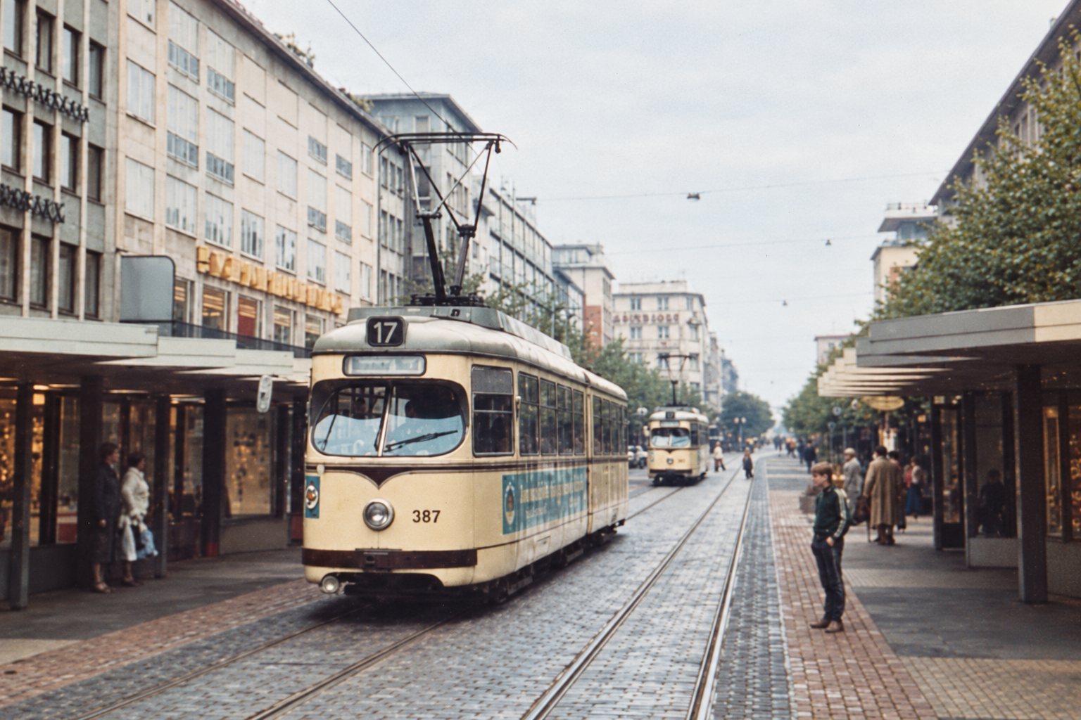 A cream-colored streetcar, number 17, on a cobblestone shopping street in Mannheim, with covered storefront arcades and pedestrians on both sides